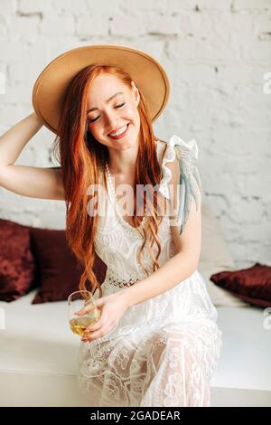Bonne jeune femme en robe boho et chapeau en dégustant du vin avec le sourire tout en étant assise sur le banc contre le mur de briques blanches Banque D'Images