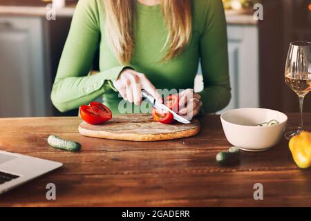 Photo rognée d'une femme blonde inconnue coupant et coupant des tomates avec un couteau tranchant dans la cuisine sur une table en bois, préparant une salade de légumes. Cuisson Banque D'Images