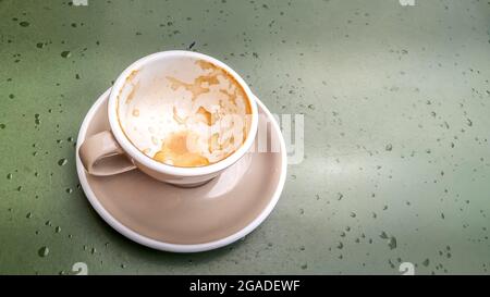 Videz la tasse de cappuccino avec le reste de mousse de lait sur la table couleur olive avec des gouttes de pluie légère. Vue de dessus, gros plan. Banque D'Images