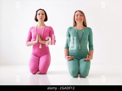 Deux jeunes femmes heureux en bonne santé dans des bodys de sport long exécutant des asanas de yoga ensemble pendant l'entraînement tout à fait méditatif ou cours de yoga de groupe, isolé Banque D'Images