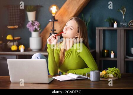 Jeune femme pensive indépendante assise à la table à la maison avec un ordinateur portable, prenant des notes, regardant attentivement dans l'air, pensant à de nouvelles idées, blo féminin Banque D'Images