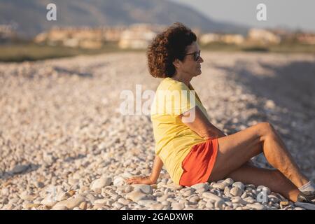 Une femme joyeuse et vitale, dans ses années 70, est assise sur une plage de galets naturelle au coucher du soleil, avec vue sur la mer Méditerranée, en Espagne Banque D'Images