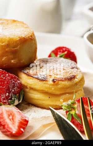 En gros plan, des crêpes au fromage cottage avec des fraises fraîches et de la confiture. Crêpes au fromage à la caillé sur une assiette blanche parsemée de sucre en poudre. Un hommage délicieux Banque D'Images