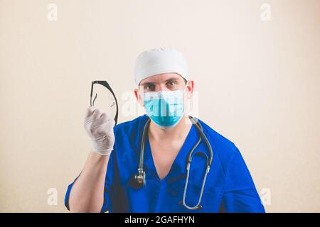 Jeune médecin en costume de chirurgien bleu et masque de protection respiratoire avec des lunettes de protection dans sa main isolée Banque D'Images