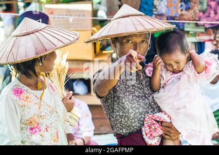 Femme birmane à la peau foncée portant un chapeau conique tenant des points pour enfants endormis à la caméra, Mine Thauk Market, Inle Lake, Myanmar
