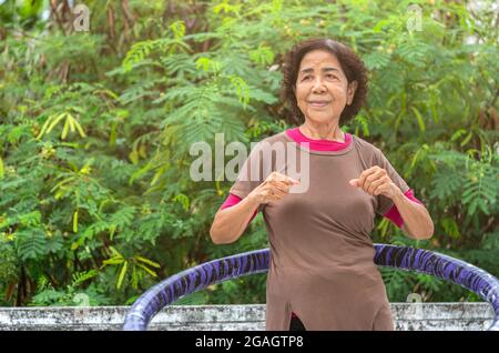 Portrait de la femme asiatique senior en bonne santé est en train de travailler par hula boucle sur la terrasse de la maison, arrière-plan partie supérieure du grand arbre, les yeux regardant la bl Banque D'Images