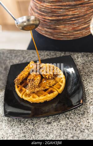 Gros plan d'une louche qui verse de la sauce ou du miel sur une assiette de poulet frit avec des céréales et des gaufres Banque D'Images