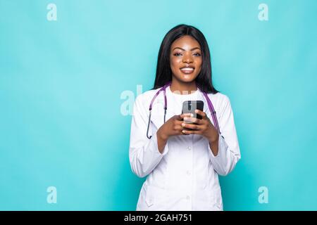 Souriante afro-américaine infirmière ou médecin femme portant un manteau médical tenant et utilisant un téléphone portable isolé sur fond turquoise Banque D'Images