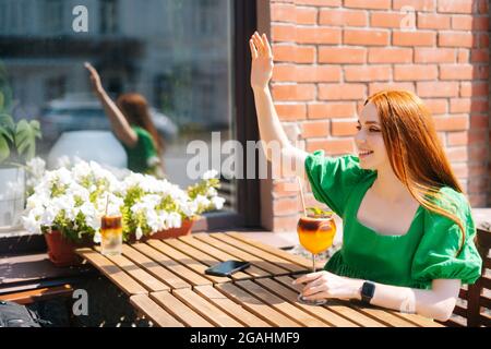 Bonne jeune femme souriante levant la main en signe de salut, saluant quelqu'un en disant bonjour avec un geste de la main assis à table dans le café extérieur Banque D'Images