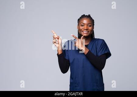 Jeune femme africaine médecin sur fond isolé gaie avec un sourire de visage pointant avec la main et le doigt vers le côté Banque D'Images