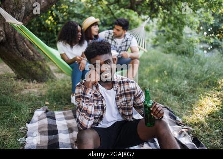 Homme noir décontracté souriant et regardant l'appareil photo tout en étant assis sur l'herbe avec une bouteille de bière dans les mains. Arrière-plan flou d'amis multiculturels bavardant, buvant et mangeant dans hamac. Pique-nique à l'extérieur. Banque D'Images