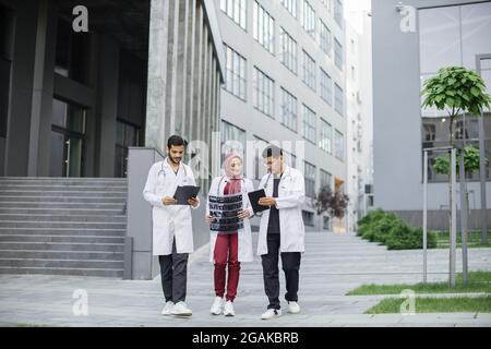 Trois médecins musulmans arabes, en costume et en manteau médicaux, discutant de la tomographie par rayons X du patient, marchant à l'extérieur sur le fond de l'hôpital moderne avec des escaliers Banque D'Images