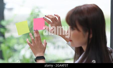 Gros plan sur une employée qui pose des notes adhésives colorées sur un mur en verre. Banque D'Images