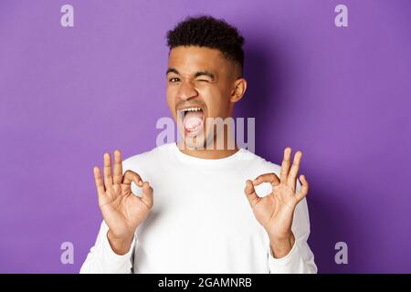 Un homme afro-américain effronté, s'assure de vous dans quelque chose, montrant des signes corrects dans l'approbation, se tenant heureux sur fond violet Banque D'Images