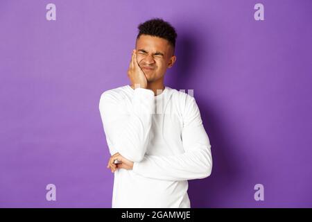 Image du jeune homme afro-américain a besoin de voir le dentiste, se plaignant de maux de dents, grimacing et toucher la joue, étant dans la douleur, debout sur le violet Banque D'Images