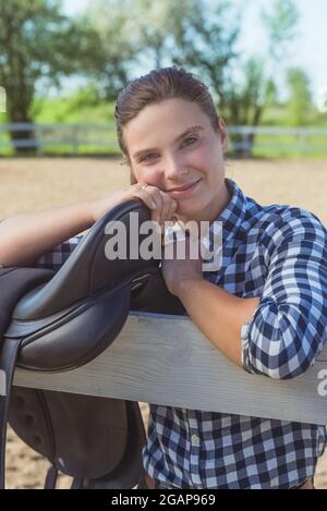 Jeune fille debout avec ses mains reposant sur la clôture en bois dans le ranch à cheval. Sourire et poser pour la caméra. Selle en cuir suspendue sur la clôture en bois au premier plan. Banque D'Images