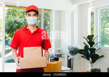 Professionnel asiatique livreur en uniforme rouge portant masque de visage tenant la boîte de cargaison et colis dans la maison, service de stockage à domicile et concept d'expédition Banque D'Images