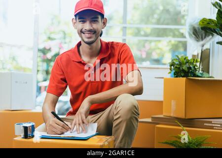 Professionnel asiatique livreur dans le sourire uniforme rouge et vérifier la boîte de cargaison et le colis dans le bureau à domicile, le service de stockage à domicile et le concept d'expédition Banque D'Images