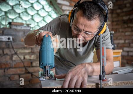 Un charpentier caucasien se concentre sur la coupe du bois avec une scie sauteuse dans son atelier. Banque D'Images