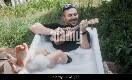 un homme joyeux dans la baignoire joue le ukulele Banque D'Images