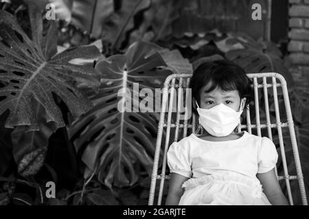 Photo en niveaux de gris d'une petite fille asiatique portant un masque facial assis sur une chaise Banque D'Images