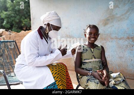 Dans cette image, une petite fille africaine courageuse sourit dans la caméra tout en recevant un vaccin par une jeune infirmière noire douce assise à côté Banque D'Images