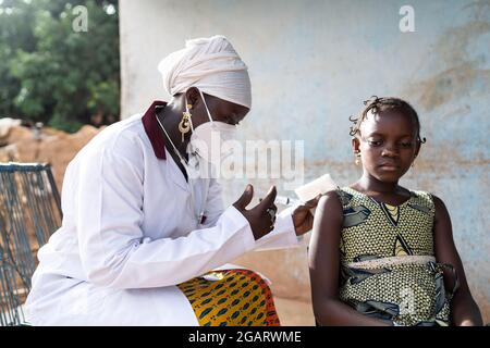 Dans cette image, une infirmière noire avec un grand masque blanc est en train d'injecter un vaccin dans le bras d'une petite fille africaine souriante tous deux assis à l'extérieur Banque D'Images