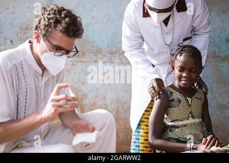 Dans cette image, un médecin caucasien masculin prépare une seringue, tandis qu'une petite fille africaine confiante en attente de son injection est rassurée par un doux Banque D'Images