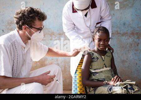 Dans cette image, un médecin caucasien masculin avec masque facial désinfecte le site d'injection sur le bras d'une petite fille africaine courageuse encouragée par une bla Banque D'Images