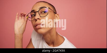 Gros plan d'une femme créative à lunettes qui regarde loin. Femme avec tête rasée sur fond rose. Banque D'Images