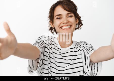 Gros plan de la belle fille moderne streth mains, atteignant pour le câlin, salutation chaleureuse, souriant heureux à l'appareil photo, debout sur fond blanc Banque D'Images