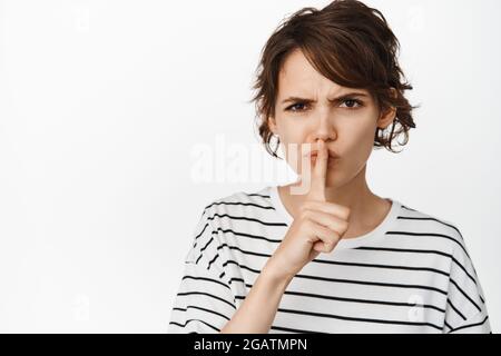 Gros plan de la fille en colère se précipitant, frognant et dire shh, appuyez le doigt sur les lèvres, geste tabou, debout contre fond blanc Banque D'Images