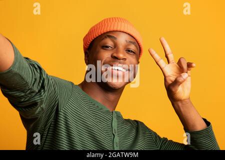 Gai Black Hipster Guy dans Orange Hat prenant Selfie et montrant le geste de paix, positif jeune Africain américain faisant autoportrait tout en vin Banque D'Images