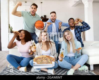 Groupe de jeunes amis internationaux avec pizza et bière regarder un match de basket-ball à la télévision, soutenant l'équipe préférée à la maison. Divers étudiants enj Banque D'Images