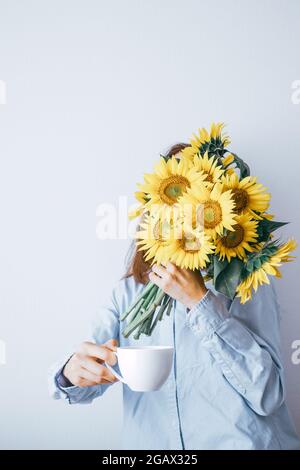 Jeune femme cachant son visage derrière un bouquet de tournesols, tenant une tasse de café sur fond blanc. . Photo de haute qualité Banque D'Images