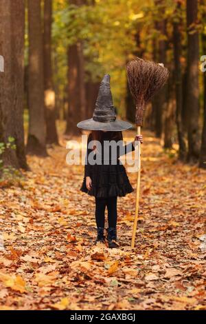 Jolie petite fille à cheveux rouges vêtue d'un costume de sorcière debout avec un balai sur fond de forêt d'automne avec espace de copie, tenant son chapeau avec la main Banque D'Images