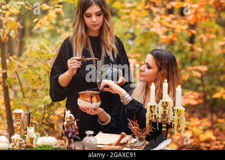 Jeunes femmes en costumes de sorcière assis à table avec des ingrédients magiques le jour d'automne dans la forêt Banque D'Images