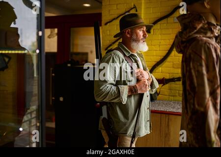 Un chasseur mâle barbu essaie de se faire un chapeau dans un magasin d'armes à feu Banque D'Images