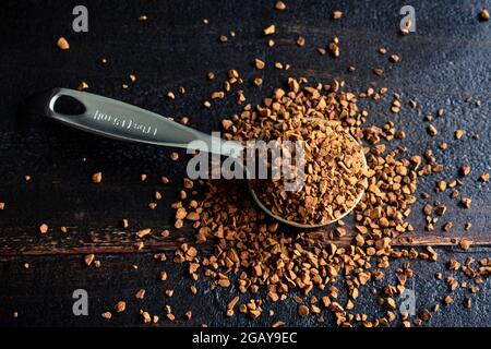 Café instantané dans une cuillère à mesurer en métal sur un fond en bois foncé : une cuillère à mesurer remplie de grains de café instantanés qui se répandent sur Banque D'Images