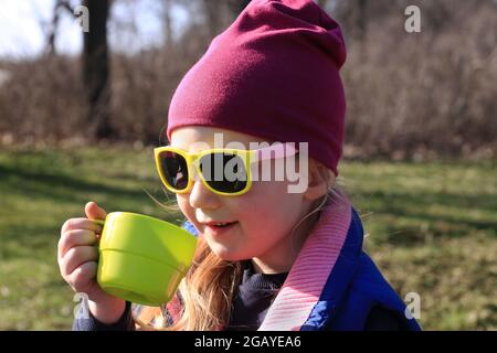 Un enfant souriant dans des vêtements colorés boit du thé dans une tasse réutilisable vert vif. Printemps nature arrière-plan. Pique-nique familial écologique. Mignonne fille dans yell Banque D'Images