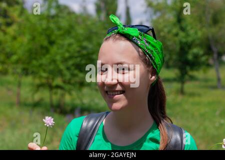 Une jeune femme émotive positive sourit, regarde la caméra et tient la fleur sauvage à la main. Portrait de jolie fille de taille basse en lunettes de soleil, chemise verte, b Banque D'Images