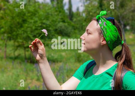 Une jeune femme émotive positive sourit, regarde la fleur sauvage qui tient dans la main. Portrait de jolie fille de taille basse en lunettes de soleil, chemise verte, banda vert Banque D'Images