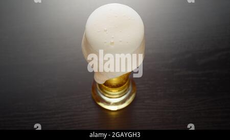 La bière dans le verre déborda. Demi-pinte de bière blonde fraîche sur la table. La mousse passe sur le bord. Vue de dessus. Banque D'Images