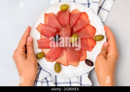 Vue ci-dessus des mains tenant une assiette blanche avec des tranches de pastrami (viande séchée des balkans) décorées d'olives Banque D'Images