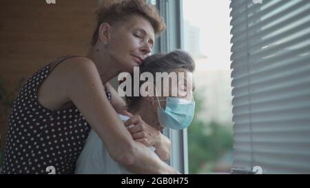 Fille adulte embrassant avec une mère aînée aimante 90 ans en masque debout à la fenêtre, des parents de différentes générations, des femmes debout Banque D'Images