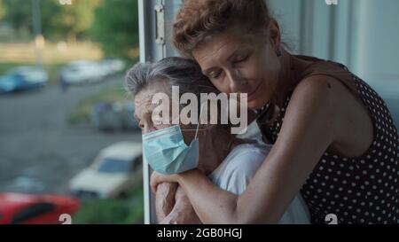 Fille adulte embrassant avec une mère aînée aimante 90 ans en masque debout à la fenêtre, des parents de différentes générations, des femmes debout Banque D'Images
