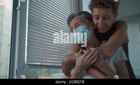 Fille mature rendant visite à une mère âgée dans une maison de soins infirmiers. Une fille âgée enferme sa mère de 90 ans dans un masque médical tout en se tenant à la fenêtre en verrouillage Banque D'Images