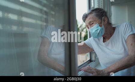 Portrait d'une femme âgée masquée à la fin des années 90 en agitant de la fenêtre d'accueil. Une femme âgée en masque de protection regarde la fenêtre et fait des vagues sur sa main Banque D'Images