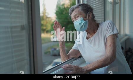 Portrait d'une femme âgée masquée à la fin des années 90 en agitant de la fenêtre d'accueil. Une femme âgée en masque de protection regarde la fenêtre et fait des vagues sur sa main Banque D'Images