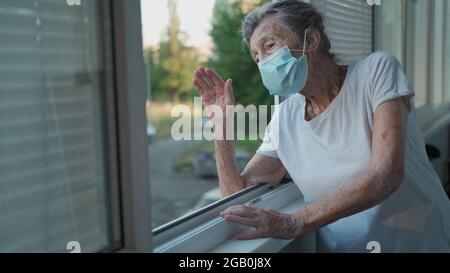 Portrait d'une femme âgée masquée à la fin des années 90 en agitant de la fenêtre d'accueil. Une femme âgée en masque de protection regarde la fenêtre et fait des vagues sur sa main Banque D'Images